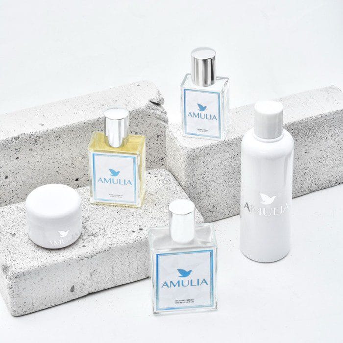 Empat botol parfum dan satu botol lotion Amulia dengan desain minimalis, berwarna putih dan transparan, diatur di atas blok-blok berwarna abu-abu.