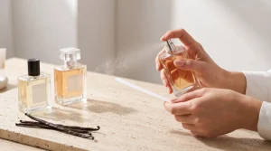 Sepasang tangan menyemprotkan parfum bernuansa vanila ke kertas uji aroma di atas meja bersama botol kaca dan biji vanila.